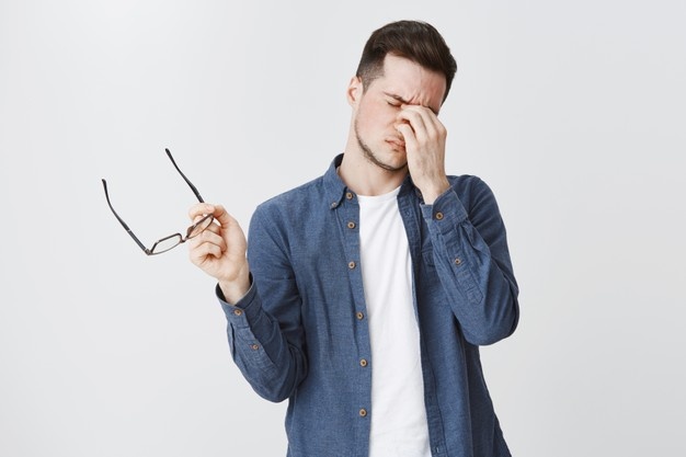 Homme en chemise bleue retirant ses lunettes et se frottant l'arête du nez, exprimant une sensation de fatigue visuelle et de tension oculaire.