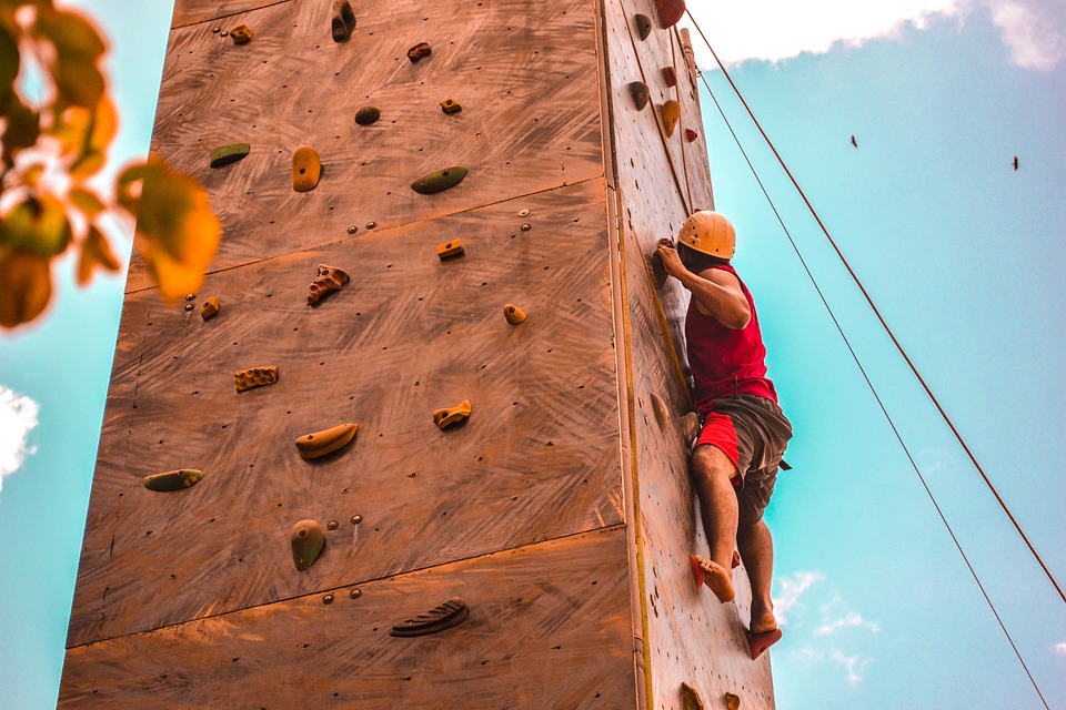 Homme en t-shirt rouge pratiquant l'escalade sur un mur artificiel en extérieur, illustrant l'effort physique et le besoin de soutien nutritionnel pour les articulations.