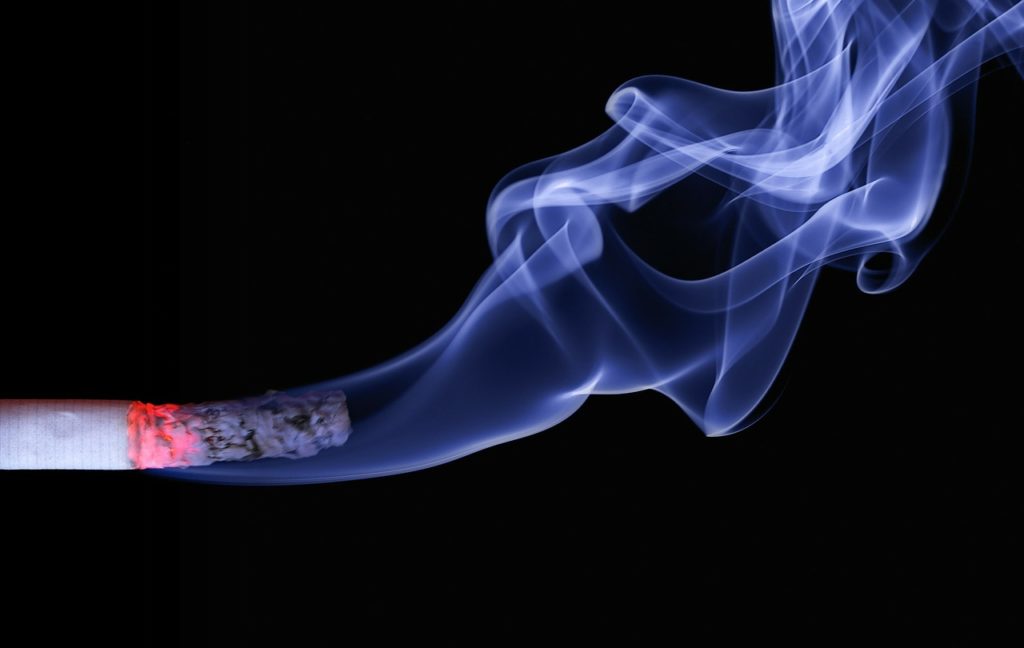 Cigarette allumée sur fond noir avec une fumée bleue épaisse, illustrant une source externe de radicaux libres et de stress oxydatif.