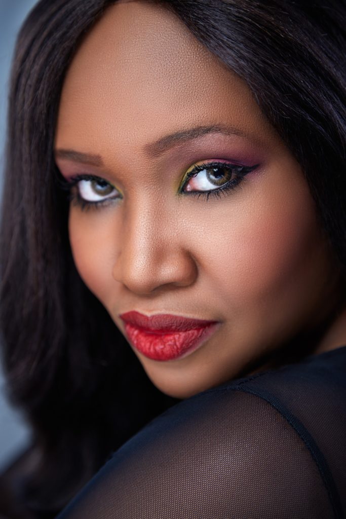 Portrait rapproché d'une femme noire ou métisse au regard intense et au maquillage précis (yeux colorés, lèvres rouges mat), symbolisant l'éclat et la mise en beauté