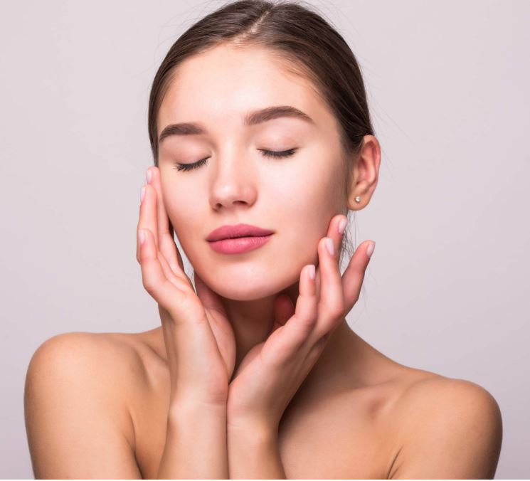 Portrait d'une jeune femme à la peau nette et lumineuse se touchant le visage avec douceur sur un fond neutre.