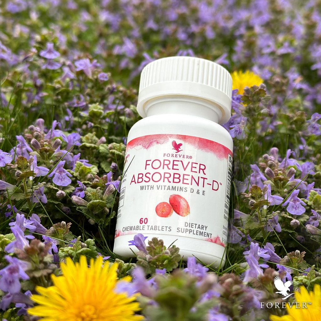 Flacon de Forever Absorbent-D posé dans un champ de fleurs violettes et de pissenlits jaunes, illustrant un complément alimentaire naturel et printanier.