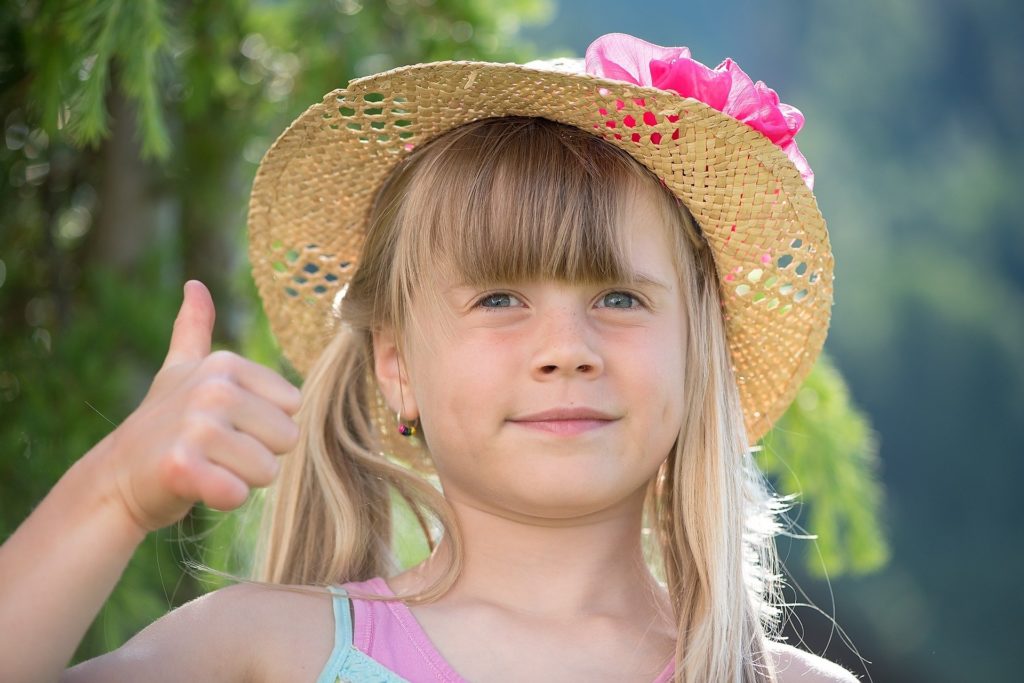 Portrait d'une petite fille souriante portant un chapeau de paille et faisant un signe de pouce levé en extérieur, illustrant la réussite d'un soin naturel.