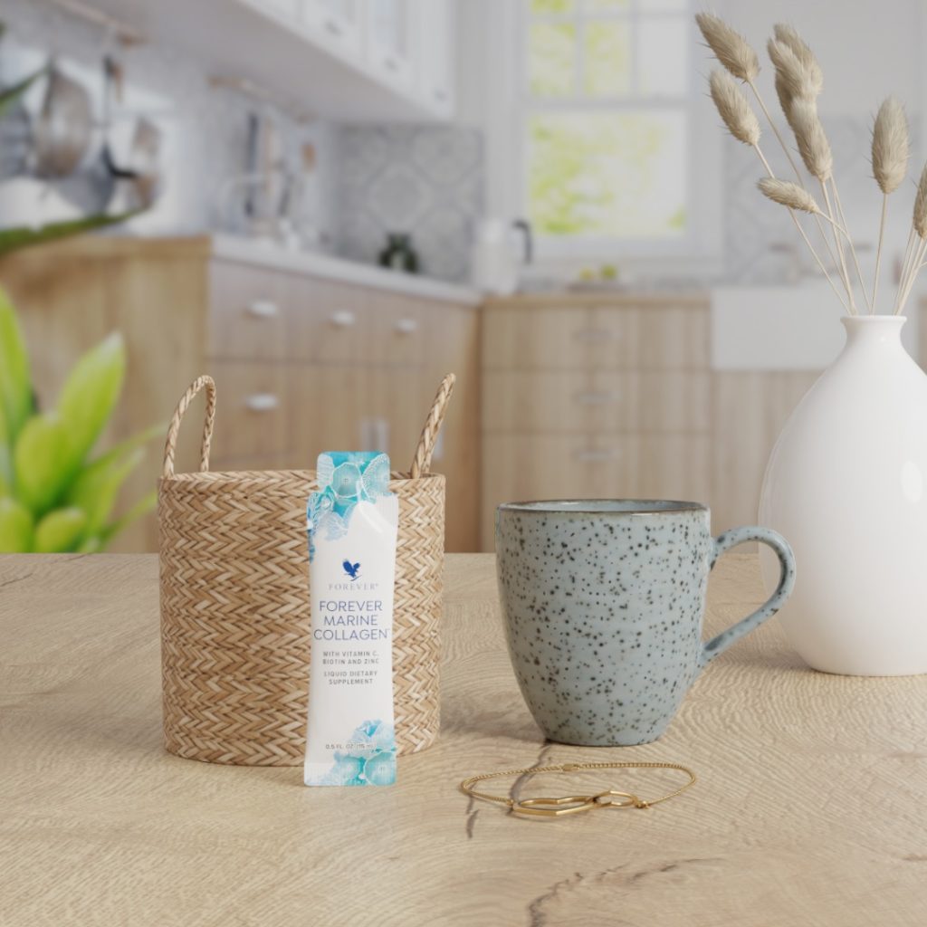Un sachet individuel de Forever Marine Collagen posé contre un petit panier en osier sur une table en bois, à côté d'une tasse et d'un vase blanc dans une cuisine lumineuse.