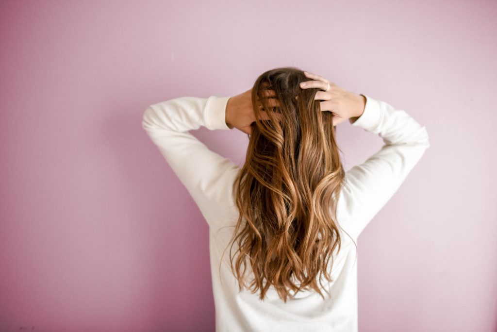 Vue de dos d'une femme aux longs cheveux châtains ondulés et brillants, passant ses mains dans sa chevelure sur un fond rose pastel.