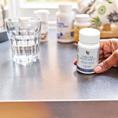 Une main tenant un flacon de Forever Multi-Maca sur une table à côté d'un verre d'eau, avec d'autres compléments alimentaires Forever en arrière-plan flou.