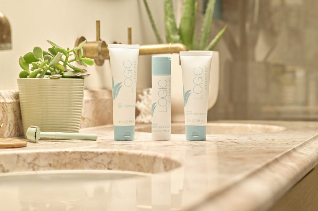 Une photo d'une salle de bain lumineuse avec un comptoir en marbre rose où sont alignés les trois produits de la gamme "Logic by Forever" : le gel nettoyant, l'essence et l'hydratant, entourés de plantes.