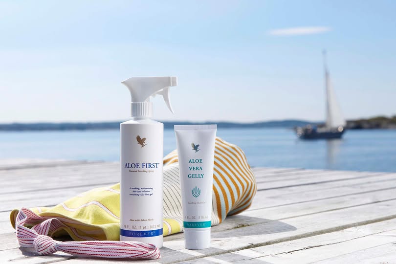Un spray Aloe First et un tube d'Aloe Vera Gelly de Forever posés sur un ponton en bois au bord de la mer, avec un voilier en arrière-plan.