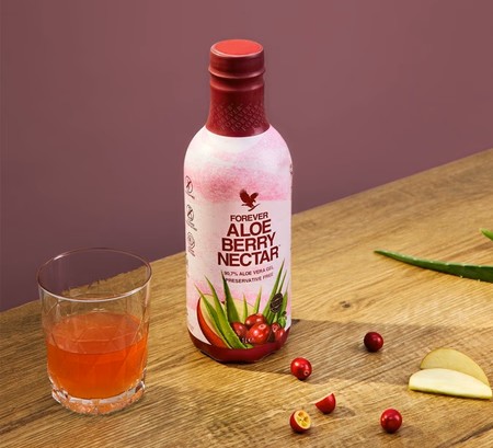 Une bouteille de Forever Aloe Berry Nectar posée sur une table en bois à côté d'un verre rempli de boisson ambrée, entourée de canneberges et de tranches de pommes.