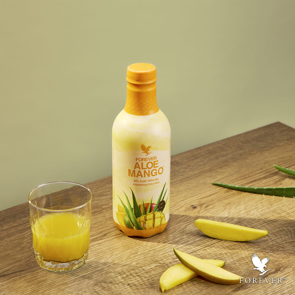 Bouteille de boisson Forever Aloe Mango sur une table en bois avec un verre rempli de jus, des tranches de mangue fraîche et des feuilles d'aloès.