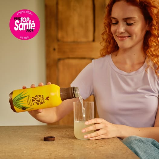 Une femme souriante aux cheveux roux verse du gel d'aloe vera Forever d'une bouteille jaune dans un verre. Un macaron rose "Prix Top Santé 2025" est visible en haut à gauche.