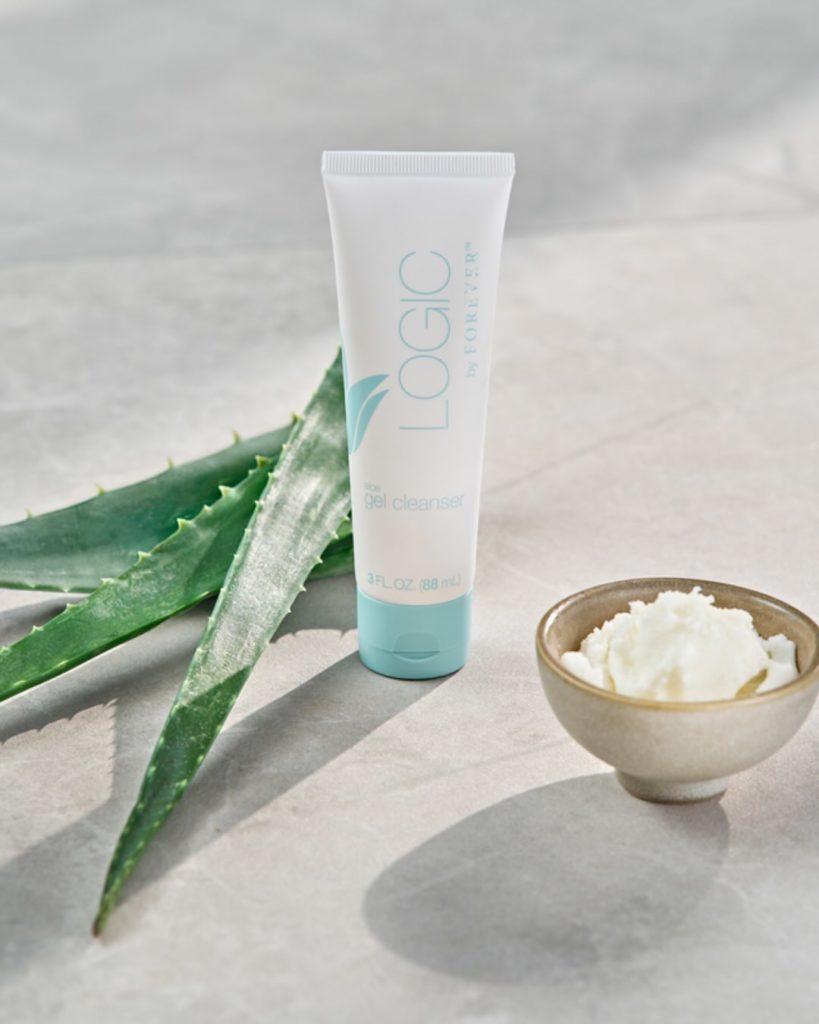 Un tube de Gel Nettoyant Visage Logic by Forever posé sur une surface grise, accompagné de feuilles d'aloe vera fraîches et d'un petit bol contenant une crème blanche.