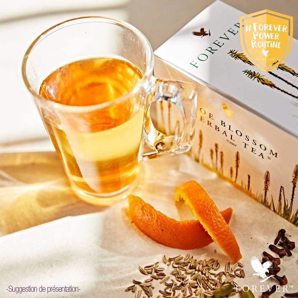 Une tasse en verre remplie d'infusion dorée à côté de sa boîte Aloe Blossom Herbal Tea, avec des zestes d'orange et des graines de fenouil.