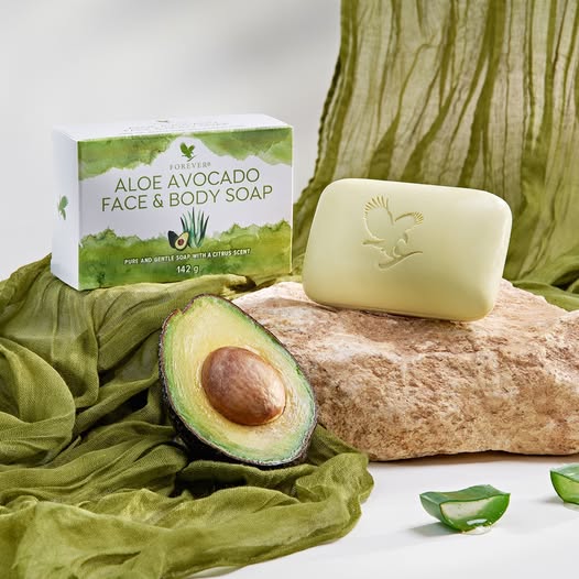 Savon Aloe Avocat Forever posé sur une pierre, à côté d'un demi-avocat frais et de morceaux d'aloe vera sur un tissu vert.