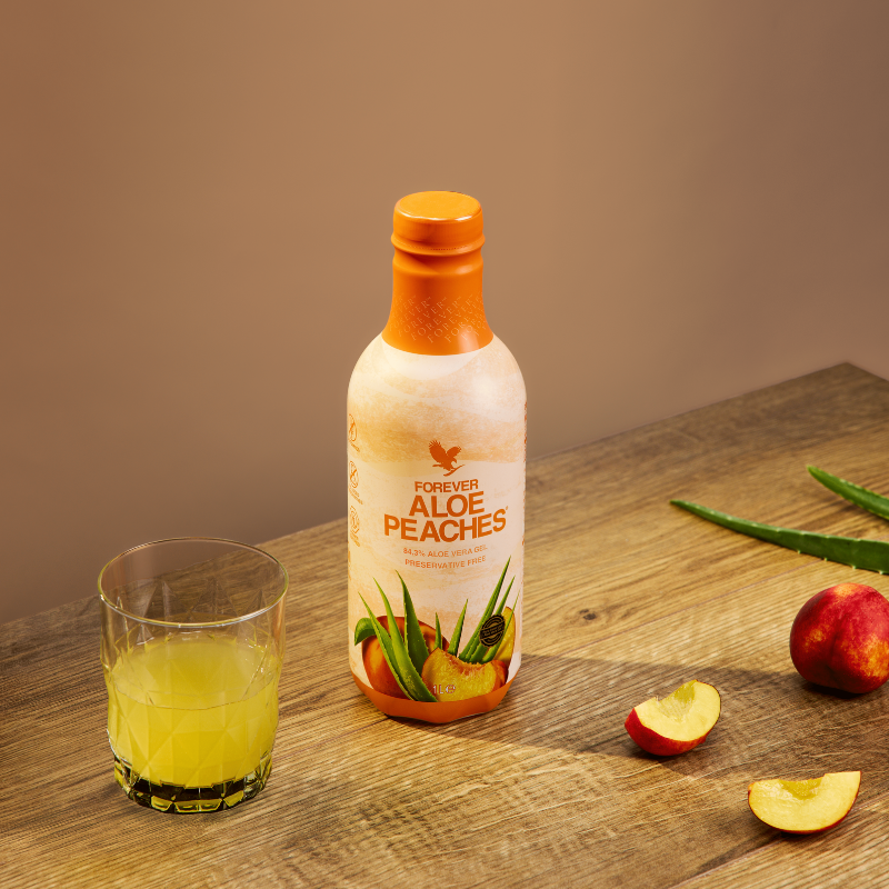 Bouteille de Forever Aloe Peaches posée sur une table en bois à côté d'un verre rempli de boisson à l'aloès, avec des quartiers de pêche fraîche et une feuille d'aloès.