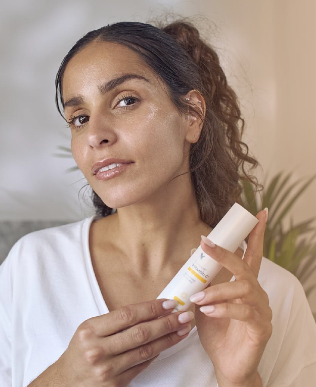 Une femme à la peau éclatante et lumineuse tenant un flacon de Forever Vitamin C , posant avec un air serein dans une pièce lumineuse.