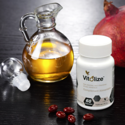 Flacon de Forever Vitolize Homme à côté d'une carafe d'huile et d'une grenade, avec quelques capsules rouges posées sur une table sombre.