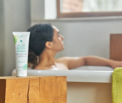 Tube d'Aloe Body Wash Forever posé sur un bloc en bois au premier plan, avec une femme se relaxant dans une baignoire en arrière-plan.