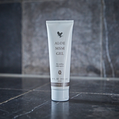 Tube blanc d'Aloe MSM Gel Forever posé verticalement sur un sol en pierre sombre devant un mur en carrelage gris.