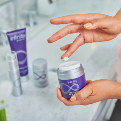 Mains tenant un pot ouvert de crème réparatrice Infinite by Forever (Restoring Crème) et prélevant une noisette de produit, avec les autres produits de la gamme en arrière-plan.