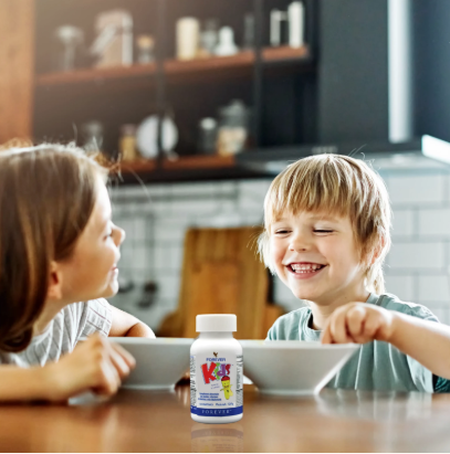 Deux jeunes enfants riant à table devant leurs bols de petit-déjeuner, avec un flacon de vitamines Forever Kids placé au centre au premier plan.