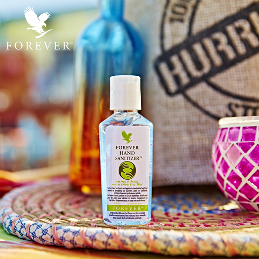 Petit flacon de Forever Hand Sanitizer posé sur un plateau en osier coloré, avec un sac en toile de jute et un bougeoir rose en arrière-plan.
