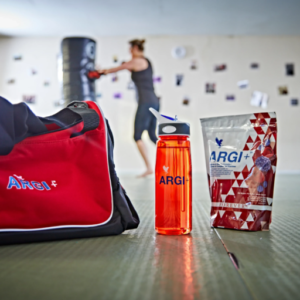 Sac de sport, gourde Argi+ et sachet de Forever ARGI Plus dans une salle de sport avec une personne s'entraînant en arrière-plan.